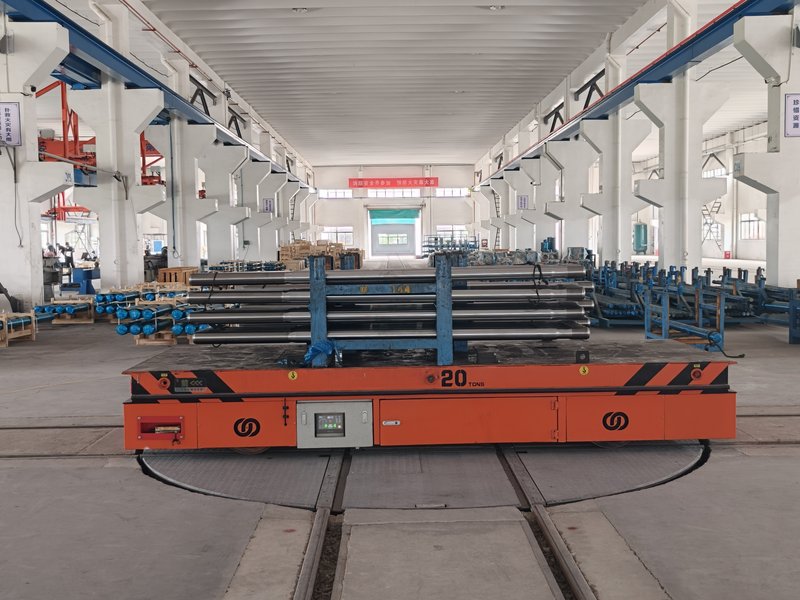 A 20-ton HENSEN heavy-duty RGV carrying long steel bar stock across an electric transfer turntable between machining workshops