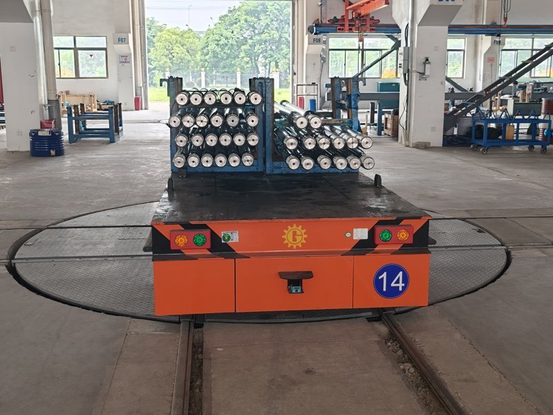 A HENSEN rail AGV positioned on a circular electric transfer turntable, carrying machined shafts between two rail lines in a heavy machining plant